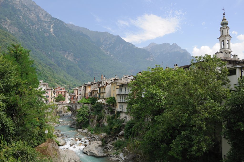 Il fiume Mera tra le case di Chiavenna, con un ponte e le montagne sullo sfondo.